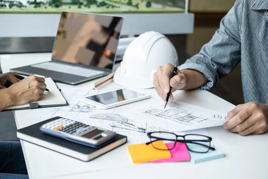 une photo montre un Diagnostiqueur Immobilier en chemise autour d'un bureau affairé à étudier des plans